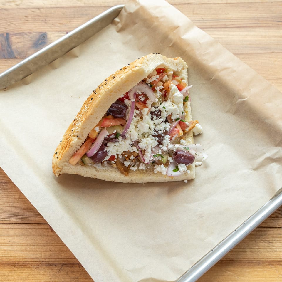 A Turkish doner kebab, stuffed with meat, feta, olives, and red onions, on parchment paper.