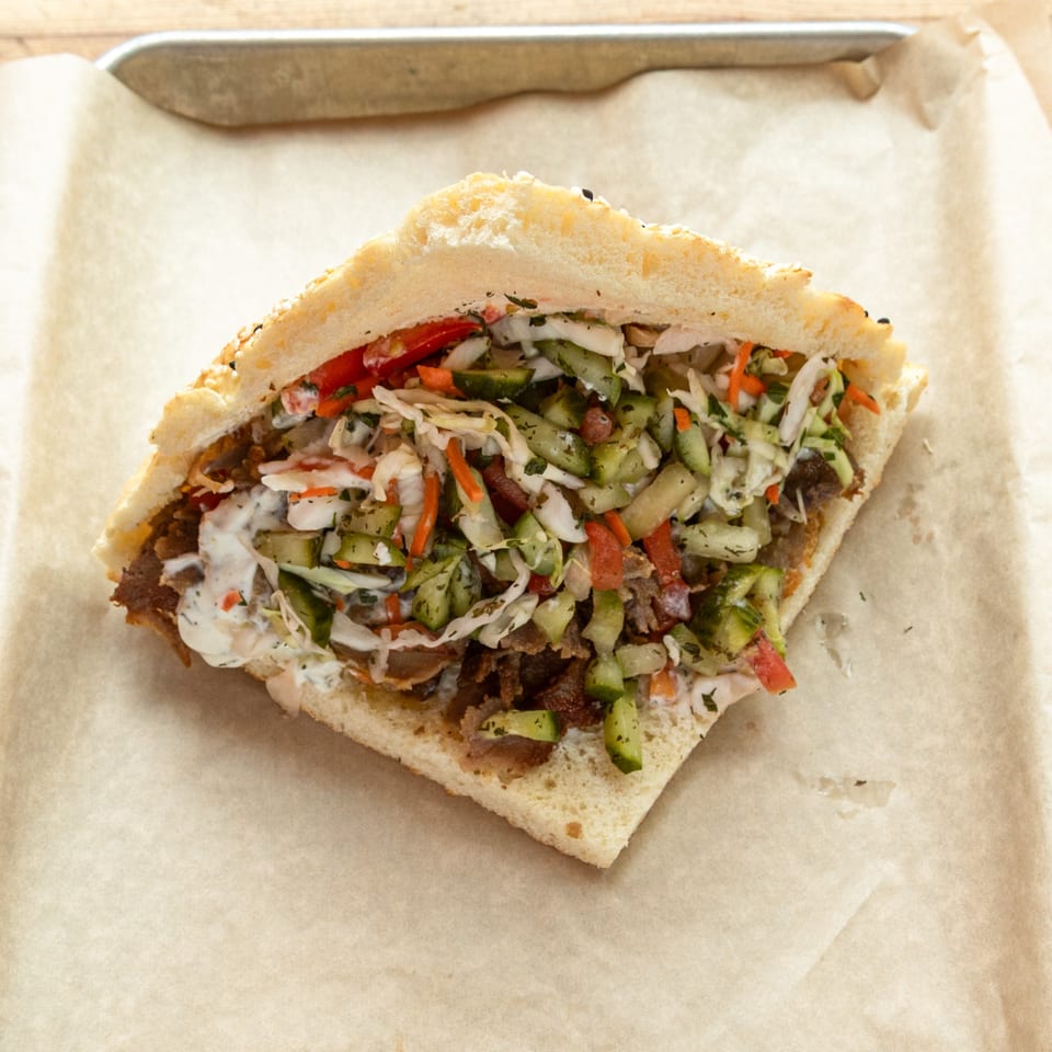 A Turkish doner kebab, filled with meat, fresh vegetables, and white sauce, on parchment paper.